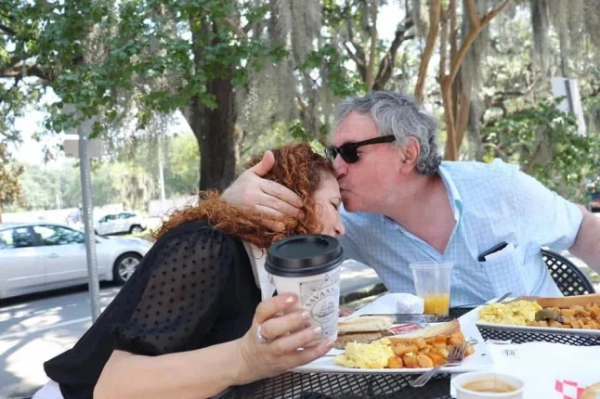 Чоловік і дружина сидять за столом на відкритому повітрі з тарілками їжі, поки чоловік цілує дружину в лоб