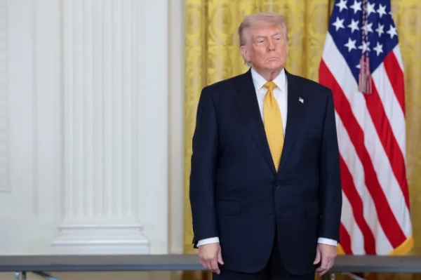 Трамп стоїть біля прапора США. Він одягнений у чорний костюм і жовту краватку. Його руки опущені вздовж тіла. Позаду нього золота завіса.