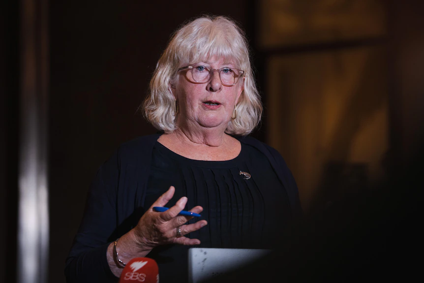 A middle-aged woman with blonde hair and glasses gesticulates