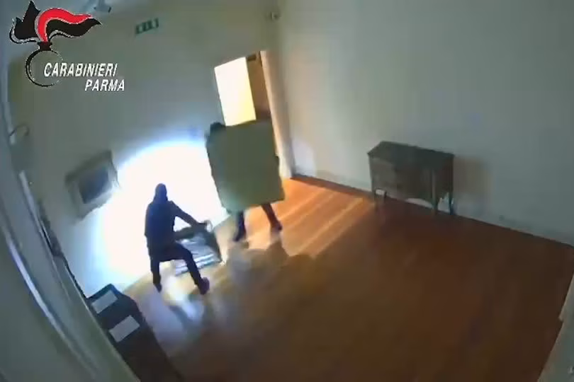 Дві фігури в масках у чорному одязі тримають картини в рамах, носячи налобні ліхтарі у великій кімнаті.