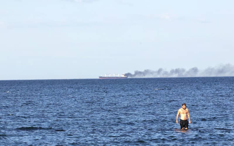 Чоловік стоїть по коліно в Чорному морі, поки чорний дим здіймається з пошкодженого російською атакою судна, Одеська область, Україна, 28 липня 2026 року. Російська армія втретє атакувала цивільне судно під прапором Ліберії, коли воно було на якорі біля підходів до Одеського порту без екіпажу чи вантажу. (Фото: Ніна Ляшонок/Ukrinform/NurPhoto) (Фото: Ніна Ляшонок / NurPhoto через AFP)