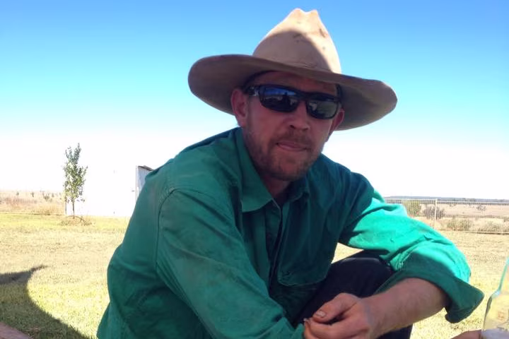 A man wearing a cowboy had and a green shirt sits on the ground holding a glass bottle
