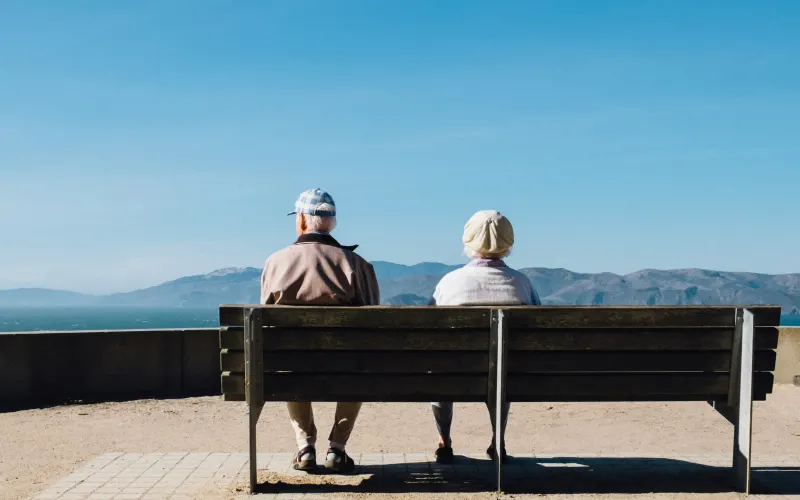 Two elderly people sit on a bench looking at a view of sea and hills.