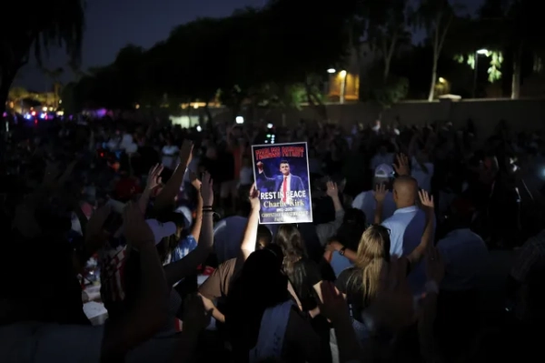 A person holds a poster with an image of slain conservative commentator Charlie Kirk as people pray next to a tribute for him at the headquarters of Turning Point USA, ahead of a memorial service for him which is to be held on September 21, in Phoenix, Arizona