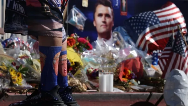 A street art memorial to Charlie Kirk with candles, flowers and a man standing in front of it wearing Trump cartoon socks